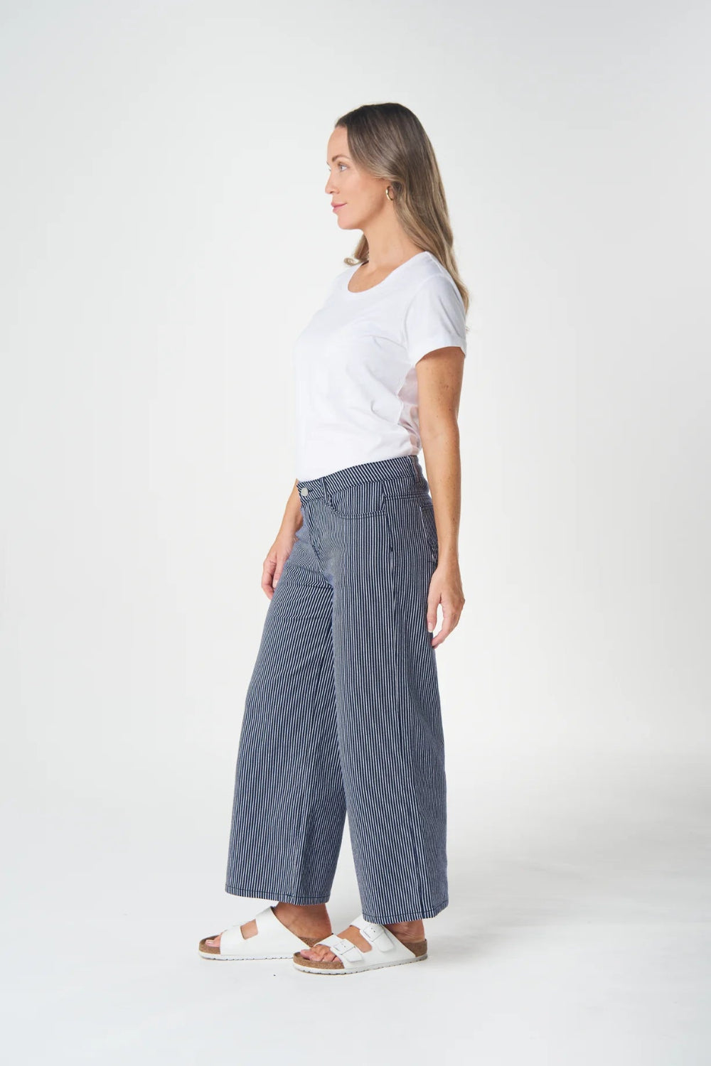 A woman shown in profile wearing a white short-sleeve T-shirt, wide-leg blue jeans, and white slide sandals against a clean white studio background.
