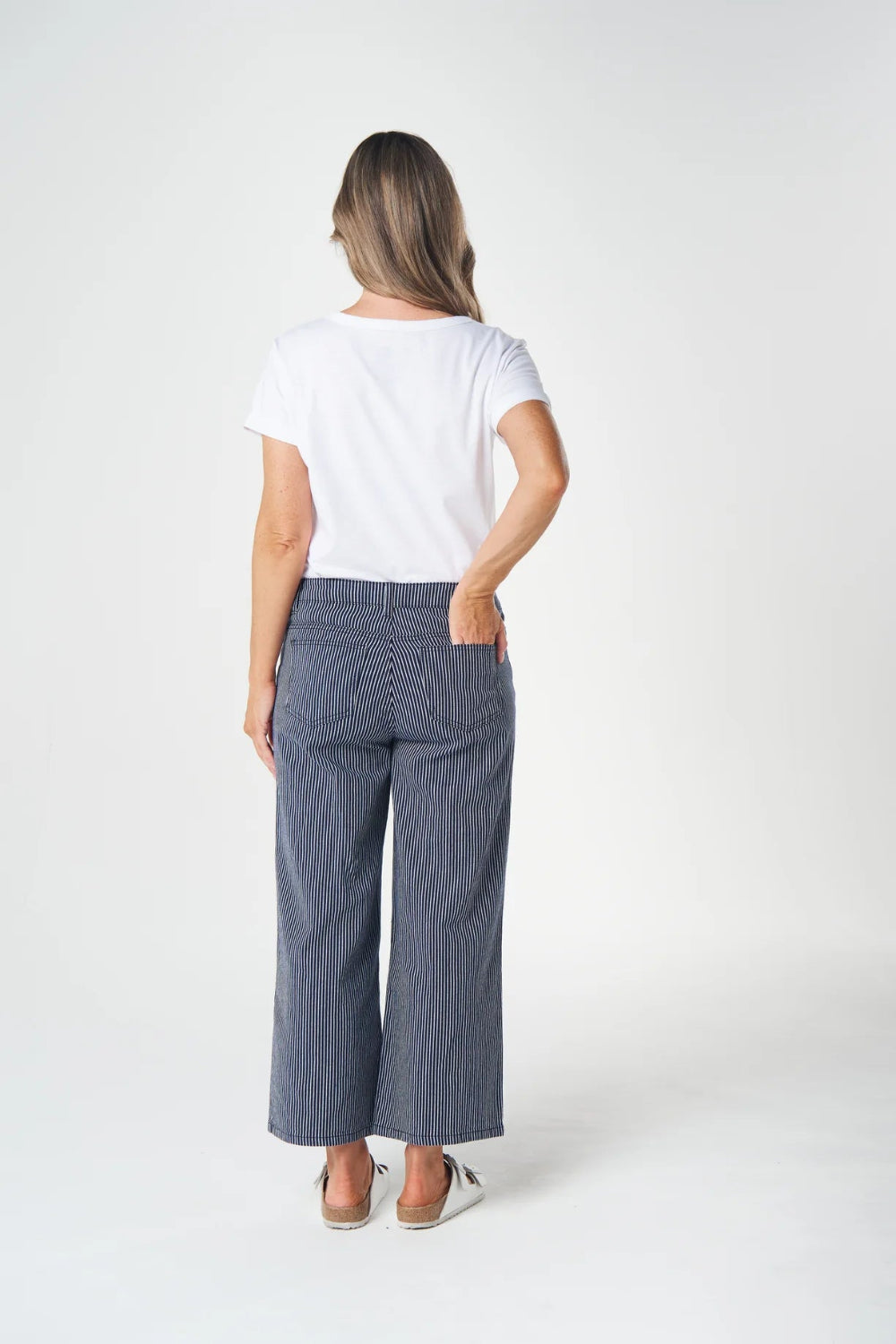 A woman is shown from behind wearing a white t-shirt and slate blue wide-leg cropped pants against a plain white studio background.