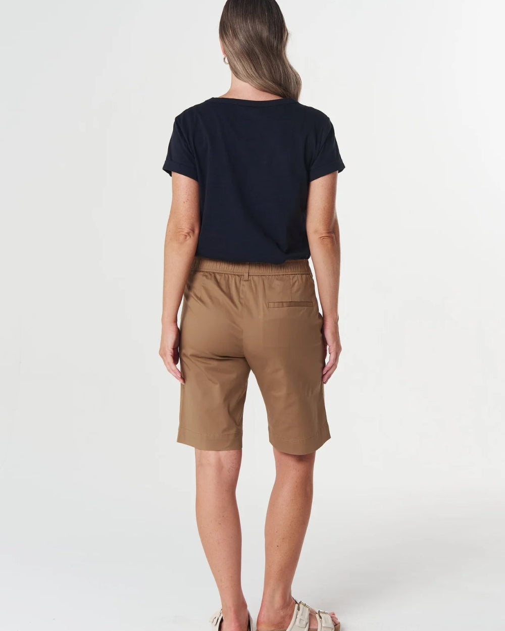 A woman shown from the back wearing a navy blue t-shirt, brown shorts, and beige sandals against a plain white studio background.