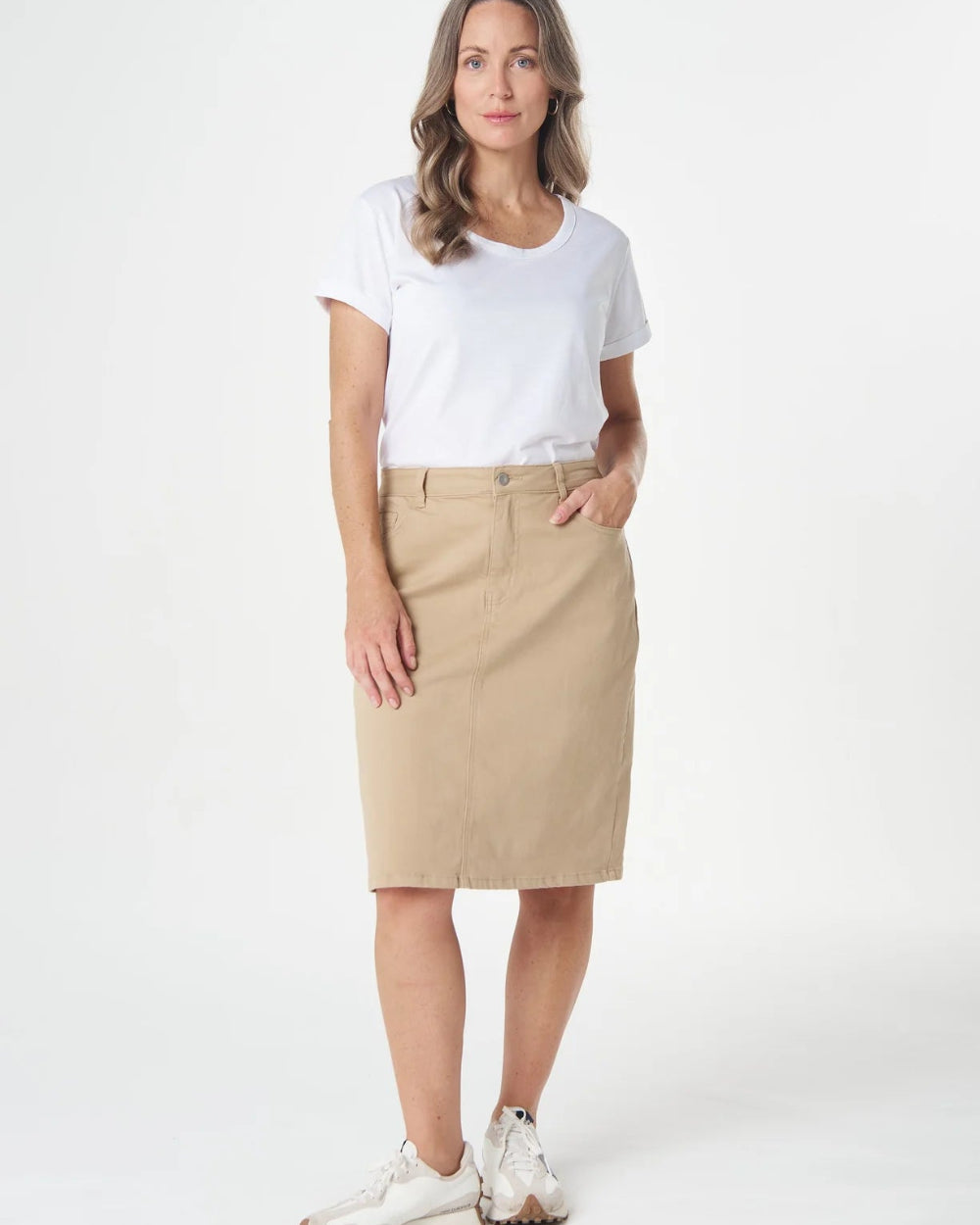 A woman modeling a beige knee-length skirt with a white t-shirt and white sneakers, standing against a plain white studio background.