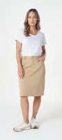 A woman modeling a beige knee-length skirt with a white t-shirt and white sneakers, standing against a plain white studio background.