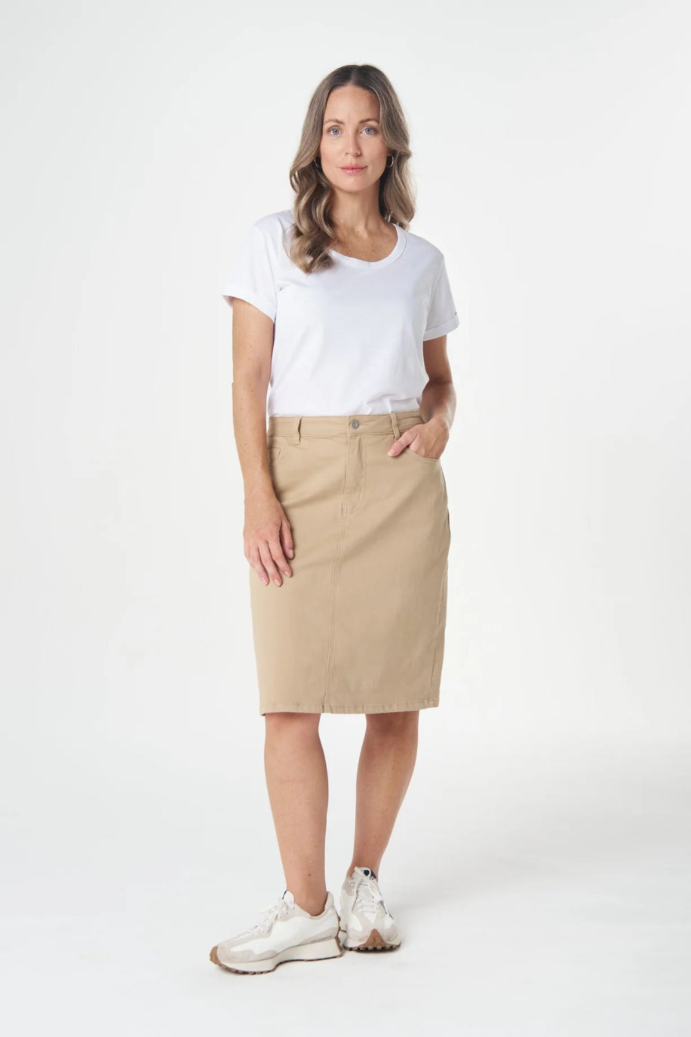 A woman modeling a beige knee-length skirt with a white t-shirt and white sneakers, standing against a plain white studio background.