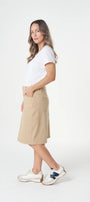 A woman modeling a beige knee-length skirt with a white t-shirt and white sneakers, standing against a plain white studio background.