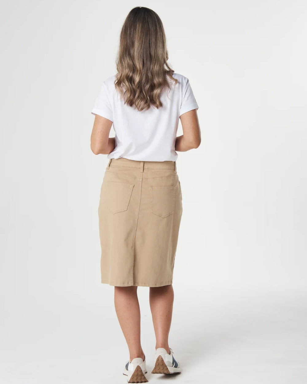 A woman viewed from behind wearing a white short-sleeve t-shirt, a beige knee-length skirt, and white sneakers with brown heels, standing against a light gray studio background.