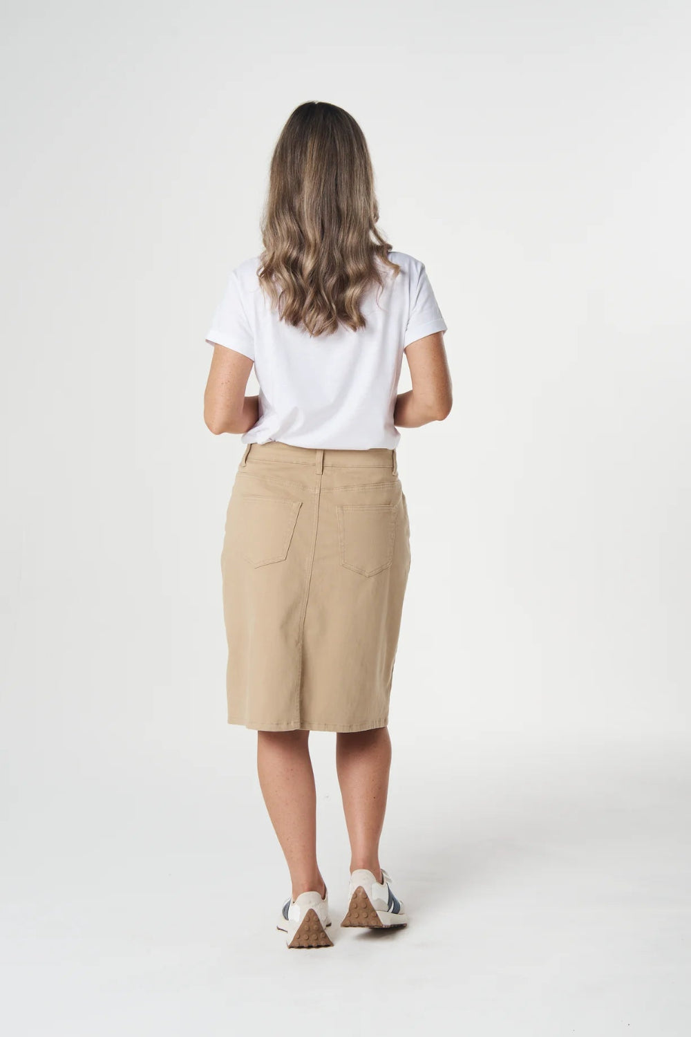 A woman viewed from behind wearing a white short-sleeve t-shirt, a beige knee-length skirt, and white sneakers with brown heels, standing against a light gray studio background.