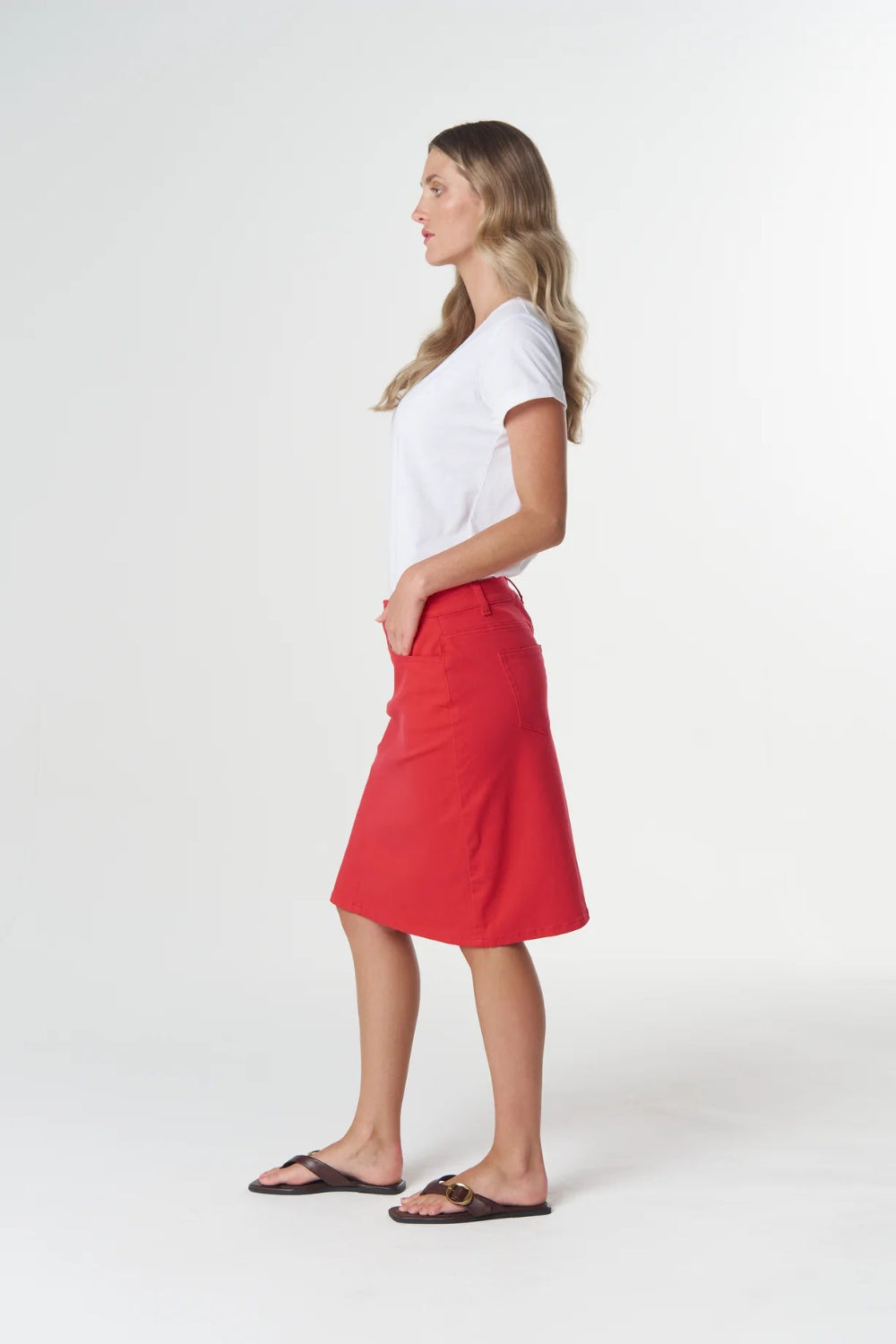 A woman in profile wearing a white short-sleeve t-shirt, a bright red A-line midi skirt, and dark brown sandals, standing against a light gray studio background.