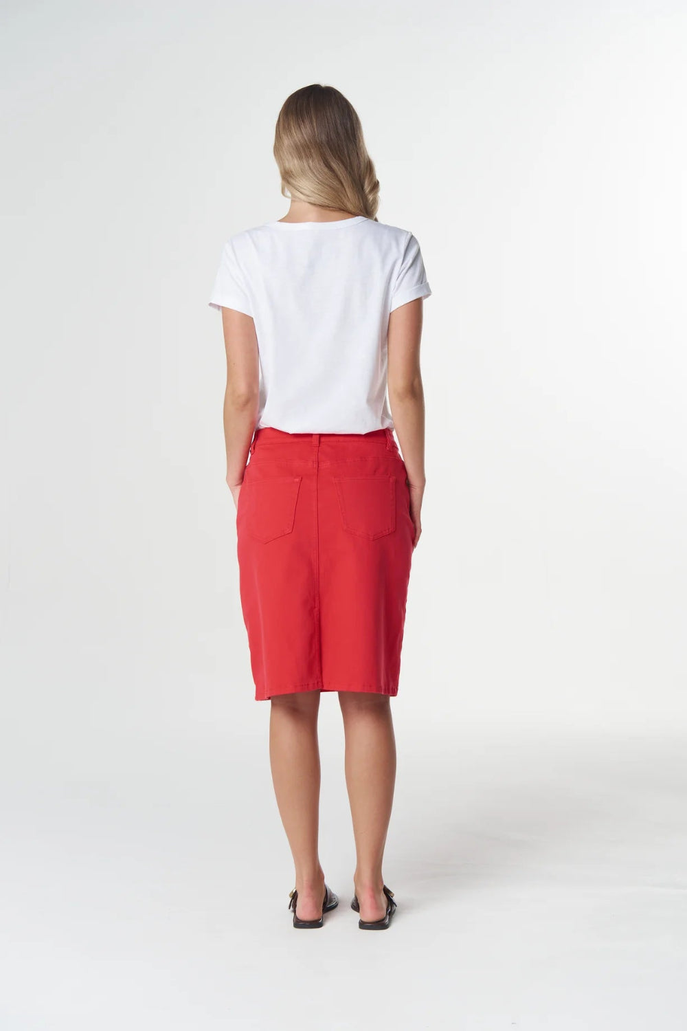 A woman viewed from behind wearing a white short-sleeve top and a bright red knee-length skirt with front patch pockets, standing against a light gray studio background.
