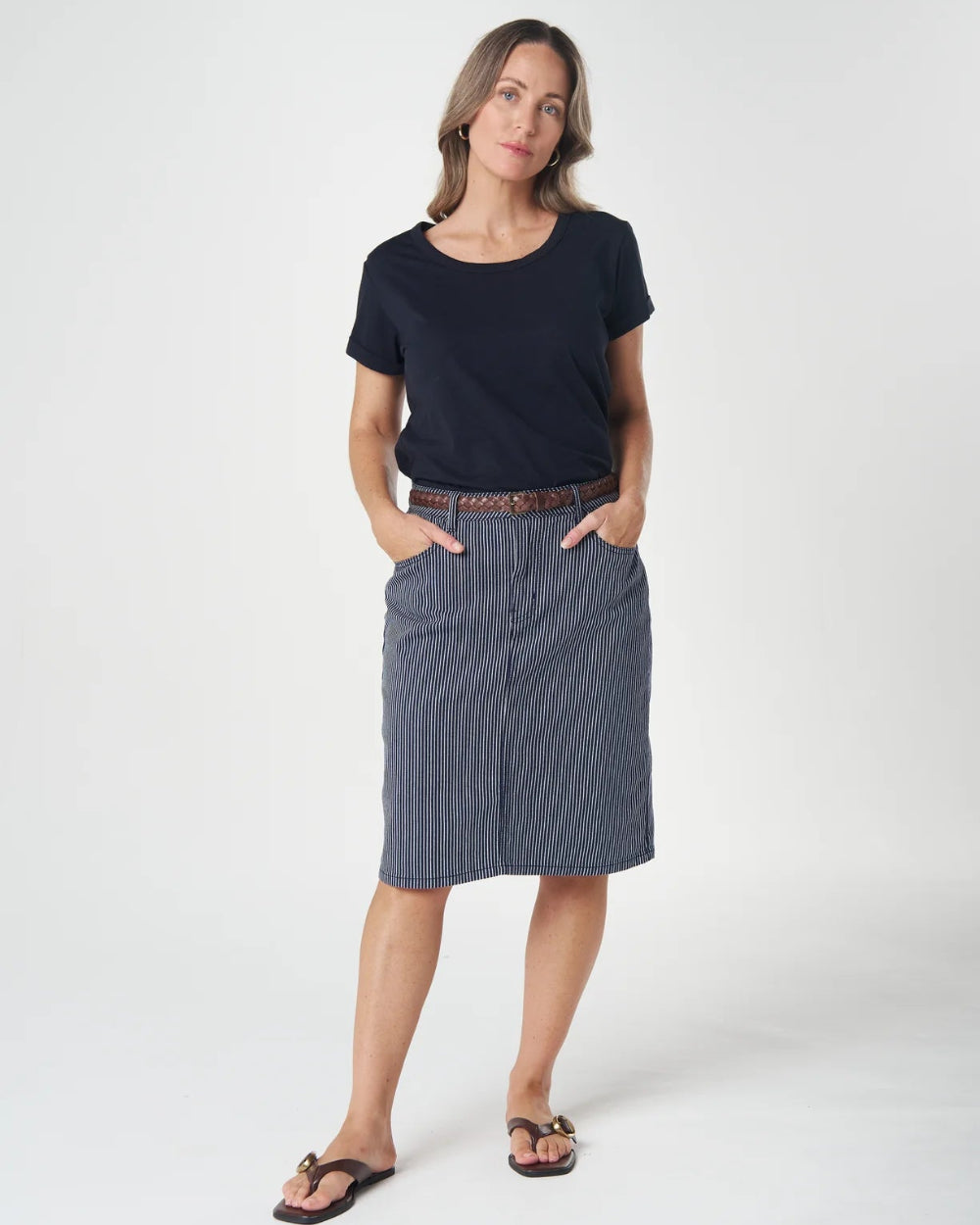 A woman modeling a dark navy blue short-sleeve t-shirt and a slate blue-gray knee-length skirt, styled with a brown belt and black sandals, against a plain white studio background.
