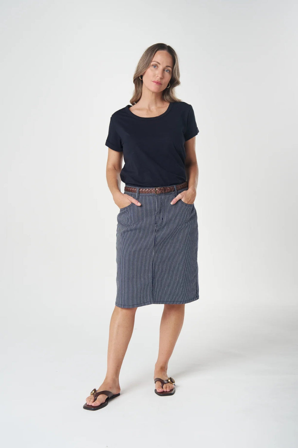 A woman modeling a dark navy blue short-sleeve t-shirt and a slate blue-gray knee-length skirt, styled with a brown belt and black sandals, against a plain white studio background.