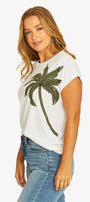 A woman wearing a white short-sleeve t-shirt with a green sequined palm tree graphic on the front, paired with blue jeans, against a plain white background.