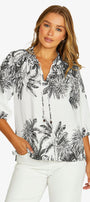 A woman wearing a white blouse with a black palm tree tropical print, paired with white pants, against a light gray studio background.