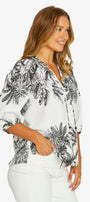 A woman wearing a white button-up blouse with a black palm leaf print, paired with white pants, shown from a side profile against a light gray studio background.