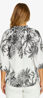 A woman viewed from the back wearing a white blouse with a black palm tree leaf pattern and white pants, set against a plain light gray background.