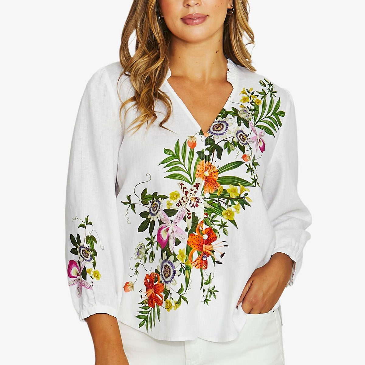 A woman wearing a white long-sleeve blouse with colorful floral embroidery on the front and sleeves, paired with white pants, against a light gray studio background.