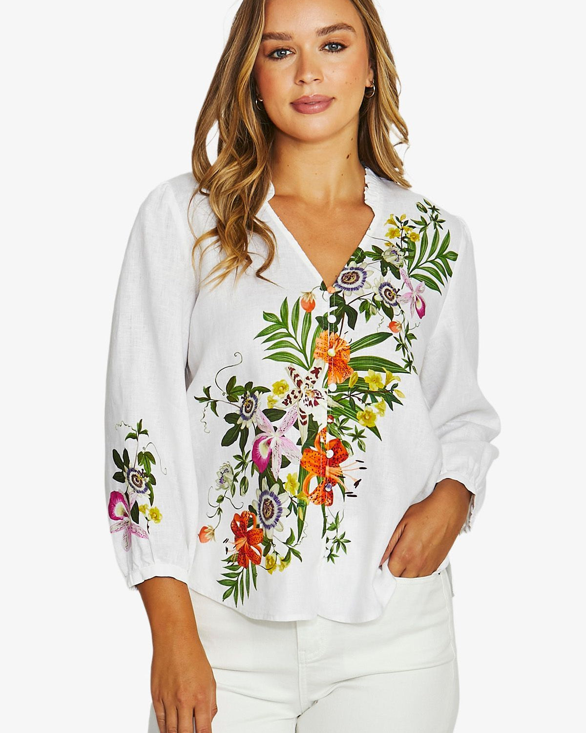 A woman wearing a white long-sleeve blouse with colorful floral embroidery on the front and sleeves, paired with white pants, against a light gray studio background.