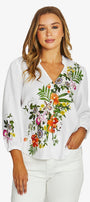A woman wearing a white long-sleeve blouse with colorful floral embroidery on the front and sleeves, paired with white pants, against a light gray studio background.
