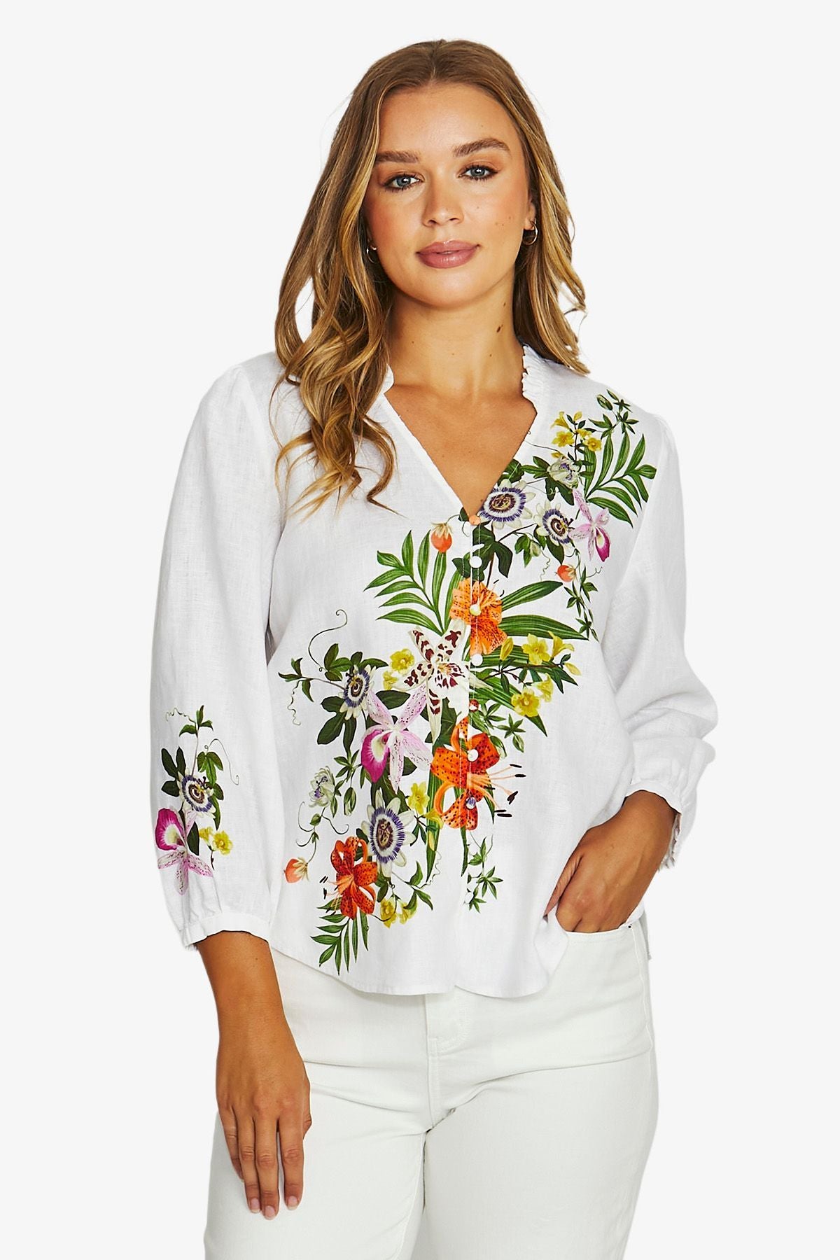 A woman wearing a white long-sleeve blouse with colorful floral embroidery on the front and sleeves, paired with white pants, against a light gray studio background.