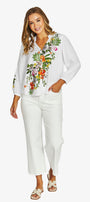 A woman modeling a white long-sleeve blouse with colorful floral embroidery on the front and white cropped pants, shown against a plain light gray studio background.