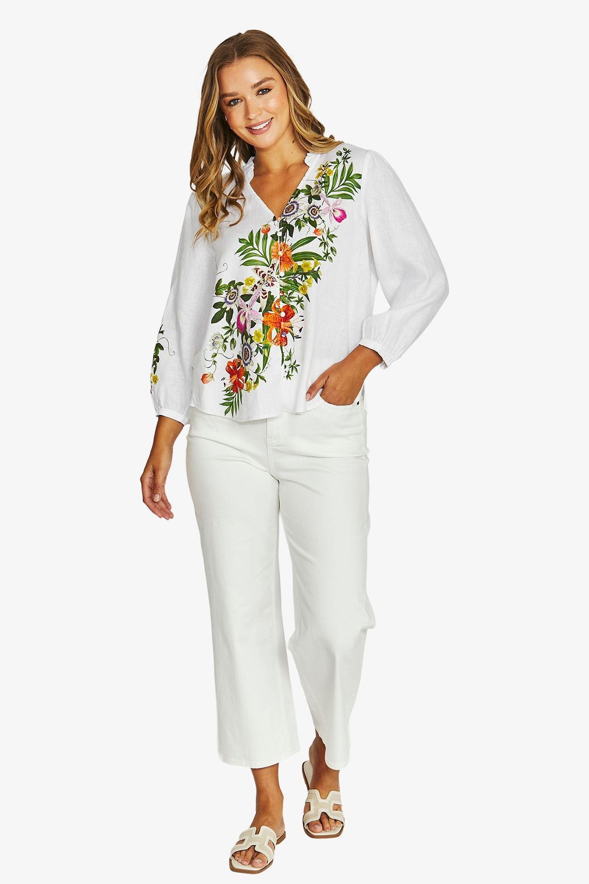 A woman modeling a white long-sleeve blouse with colorful floral embroidery on the front and white cropped pants, shown against a plain light gray studio background.