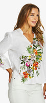 A woman wearing a white long-sleeve blouse with colorful floral embroidery on the front and sleeve, paired with white pants, against a light gray studio background.