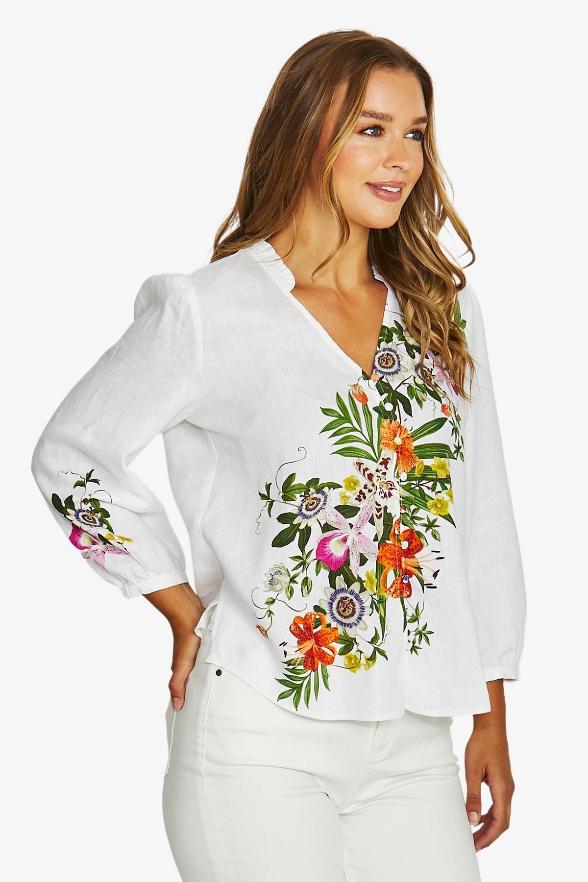 A woman wearing a white long-sleeve blouse with colorful floral embroidery on the front and sleeve, paired with white pants, against a light gray studio background.