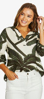 A woman wearing a long-sleeve wrap-style blouse with a dark green palm leaf pattern on an off-white background, paired with white pants, against a light gray studio background.
