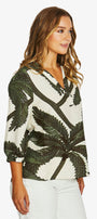 A woman wearing a white long-sleeve blouse with a dark green palm tree leaf print, paired with white pants, photographed against a light gray studio background.