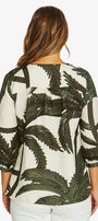 A woman viewed from the back wearing a cream-colored blouse with a dark green palm leaf pattern and white pants, set against a plain light gray background.