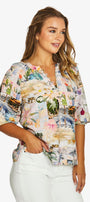 Front view  a short sleeve V-neck blouse with a colorful tropical and scenic patchwork print, paired with white pants. 