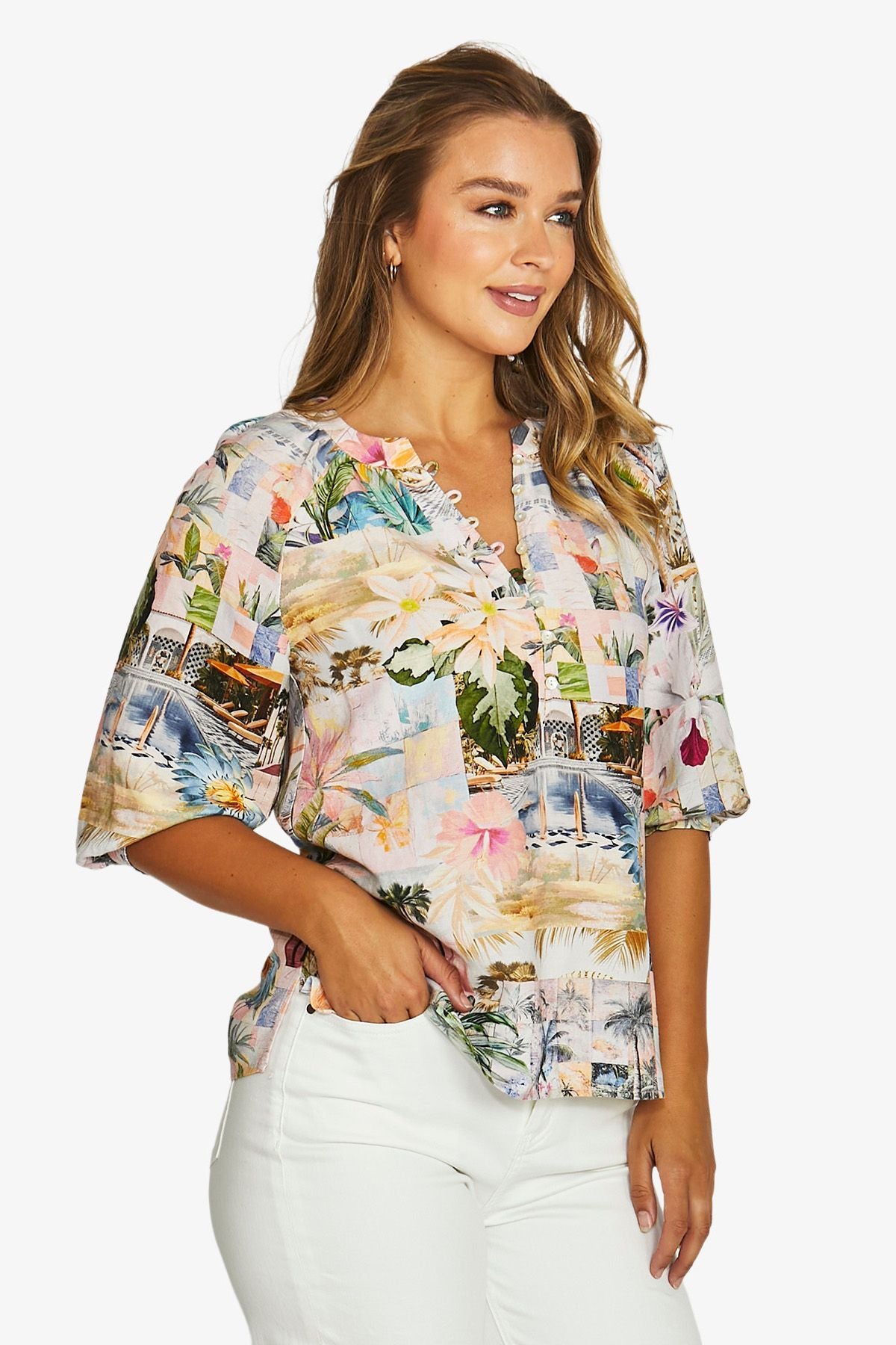 Front view  a short sleeve V-neck blouse with a colorful tropical and scenic patchwork print, paired with white pants. 