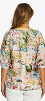  back  view wearing a short sleeve top with a colorful tropical and beach themed print, paired with white pants.