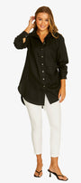 A woman modeling a black long-sleeve button-up shirt with white pants and black sandals, standing against a light gray studio background.
