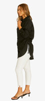 A woman shown in profile wearing a loose black long-sleeve top with a side tie detail, white skinny pants, and black heeled sandals against a plain light gray background.
