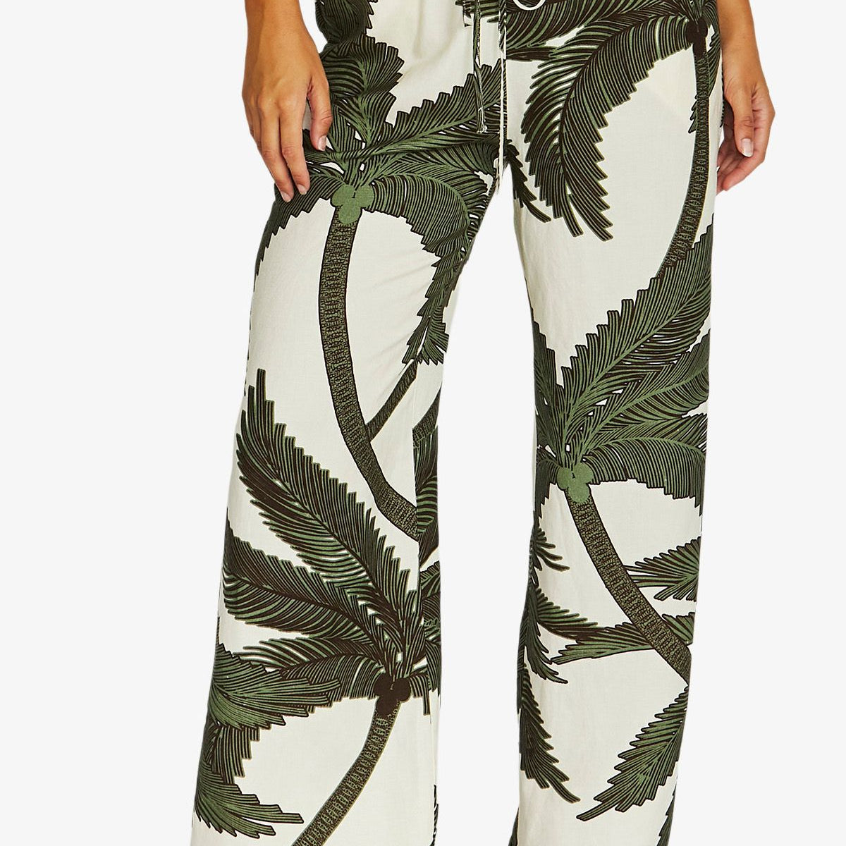 A person wearing wide-leg drawstring pants with an off-white background and a dark green palm tree print, paired with a white top and tan heeled sandals against a light gray studio background.