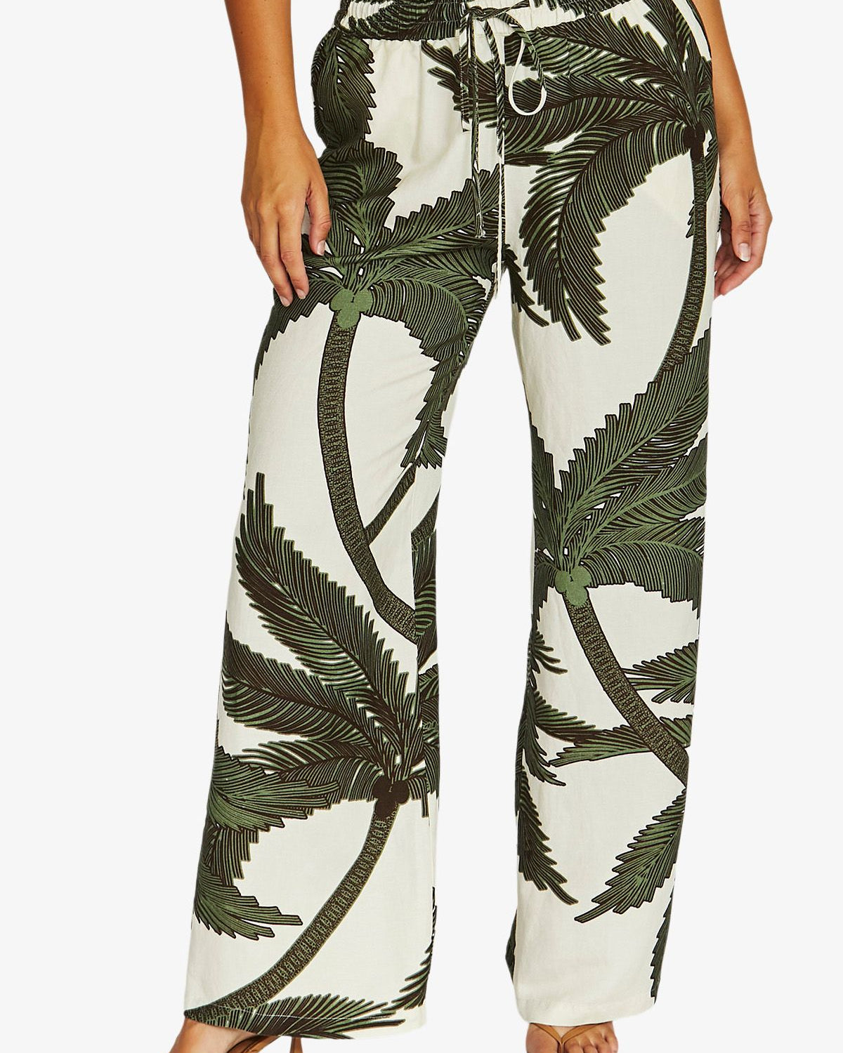 A person wearing wide-leg drawstring pants with an off-white background and a dark green palm tree print, paired with a white top and tan heeled sandals against a light gray studio background.