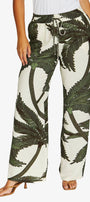 A person wearing wide-leg drawstring pants with an off-white background and a dark green palm tree print, paired with a white top and tan heeled sandals against a light gray studio background.