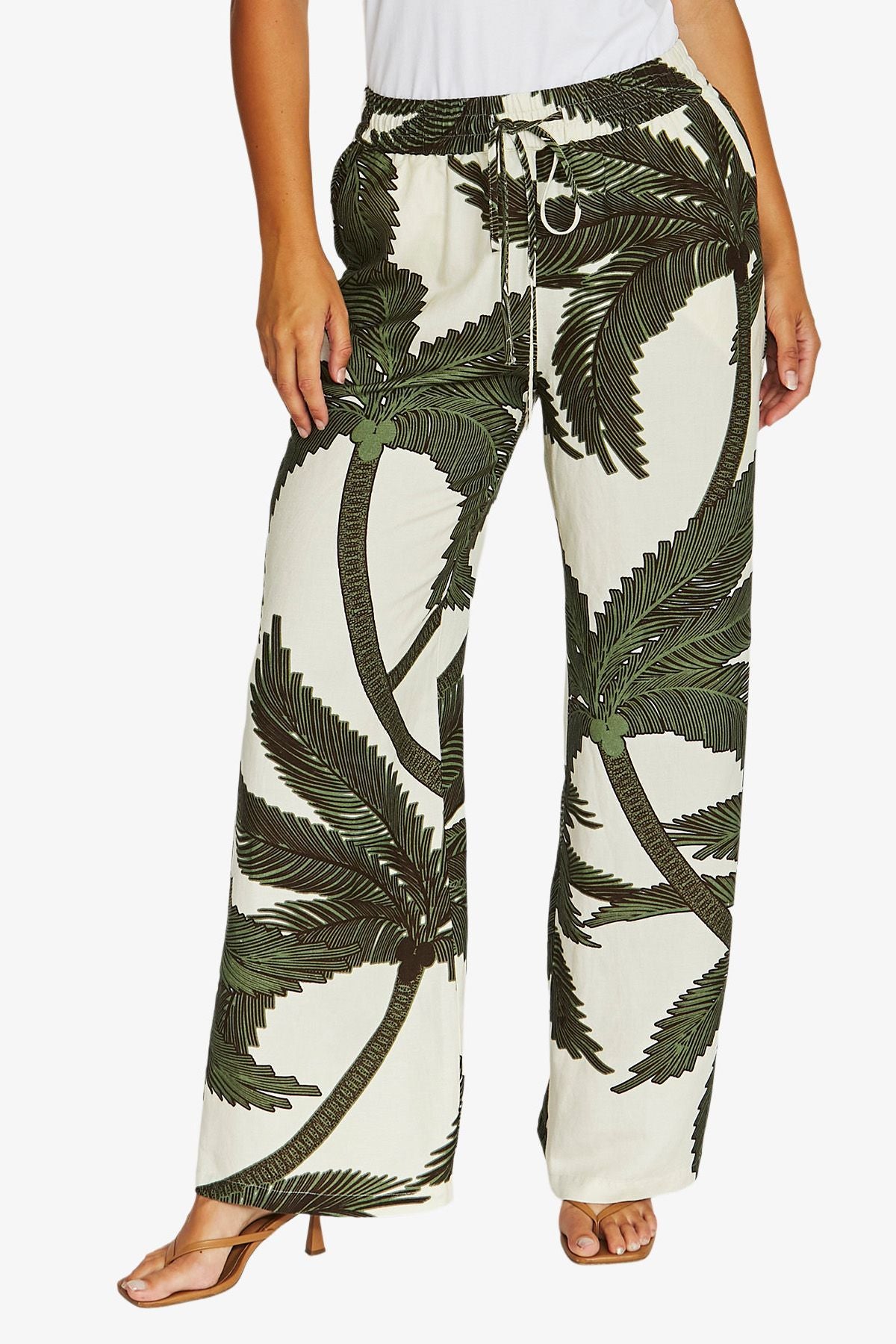 A person wearing wide-leg drawstring pants with an off-white background and a dark green palm tree print, paired with a white top and tan heeled sandals against a light gray studio background.