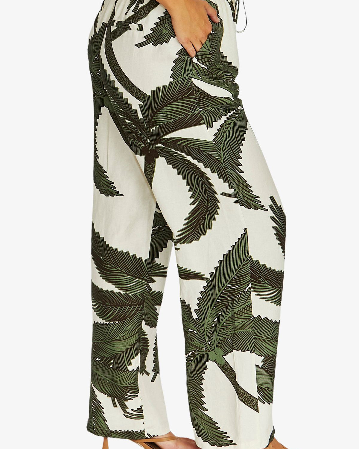 A person wearing high-waisted wide-leg pants with a bold green palm leaf print on an off-white background, paired with tan heeled sandals against a light gray studio background.