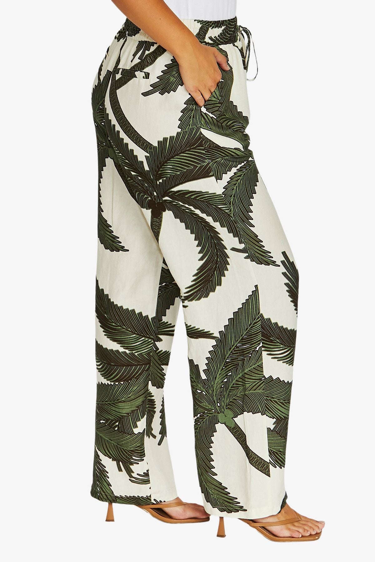A person wearing high-waisted wide-leg pants with a bold green palm leaf print on an off-white background, paired with tan heeled sandals against a light gray studio background.