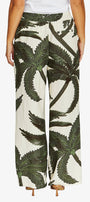 A person wearing wide-leg pants with an allover dark green palm tree print on an off-white background, paired with a white top and tan heels, shown against a light gray studio background.
