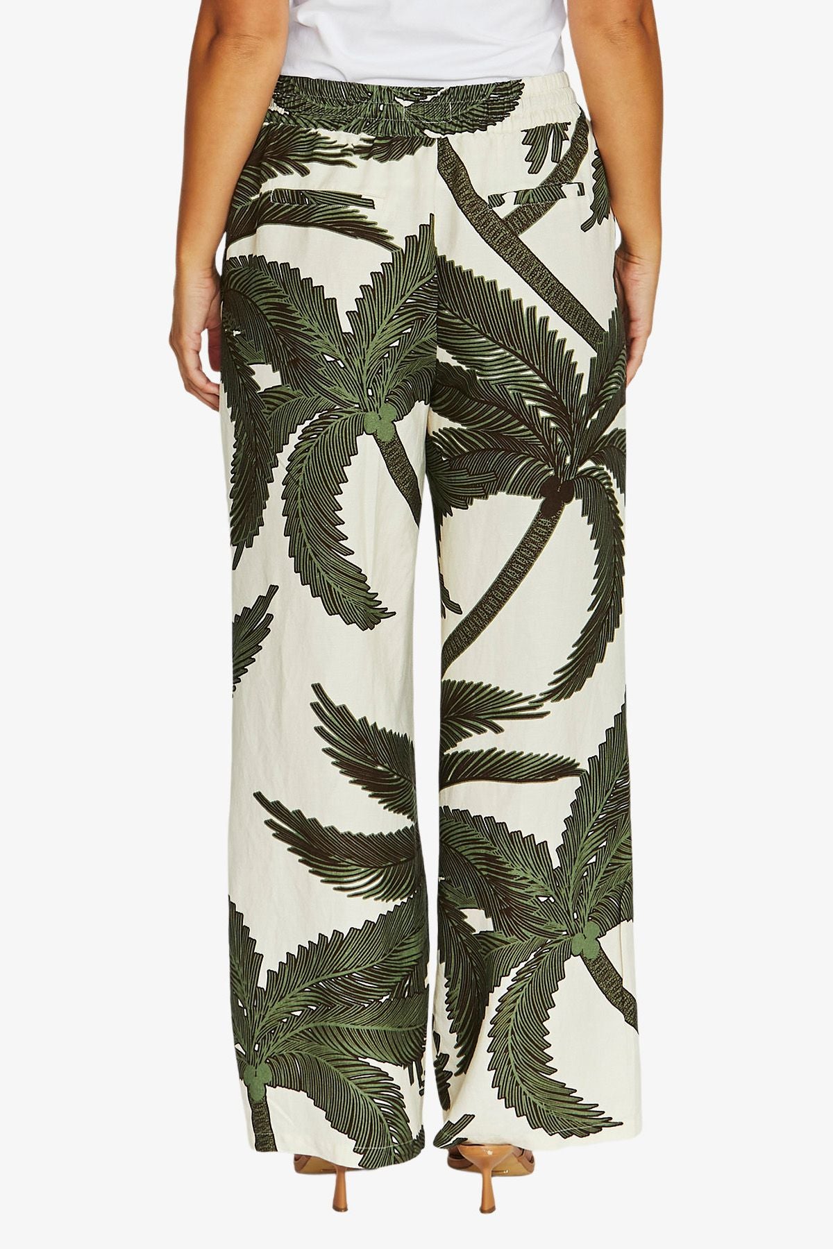 A person wearing wide-leg pants with an allover dark green palm tree print on an off-white background, paired with a white top and tan heels, shown against a light gray studio background.