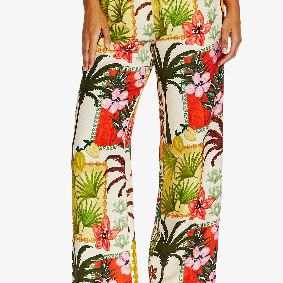 Front View high waisted, wide leg trousers with a vibrant tropical patchwork print featuring palm trees, hibiscus flowers, and red orange panels on a cream background. The pants are styled with a matching belt and brown heeled sandals.