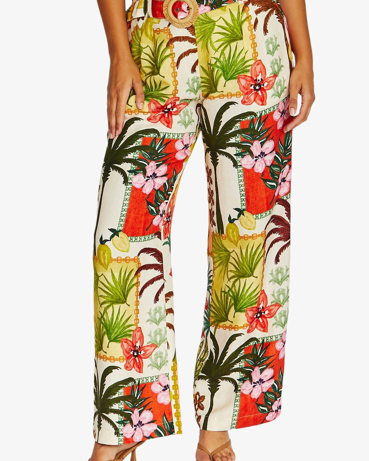 Front View high waisted, wide leg trousers with a vibrant tropical patchwork print featuring palm trees, hibiscus flowers, and red orange panels on a cream background. The pants are styled with a matching belt and brown heeled sandals.
