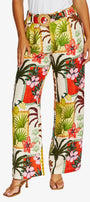 Front View high waisted, wide leg trousers with a vibrant tropical patchwork print featuring palm trees, hibiscus flowers, and red orange panels on a cream background. The pants are styled with a matching belt and brown heeled sandals.