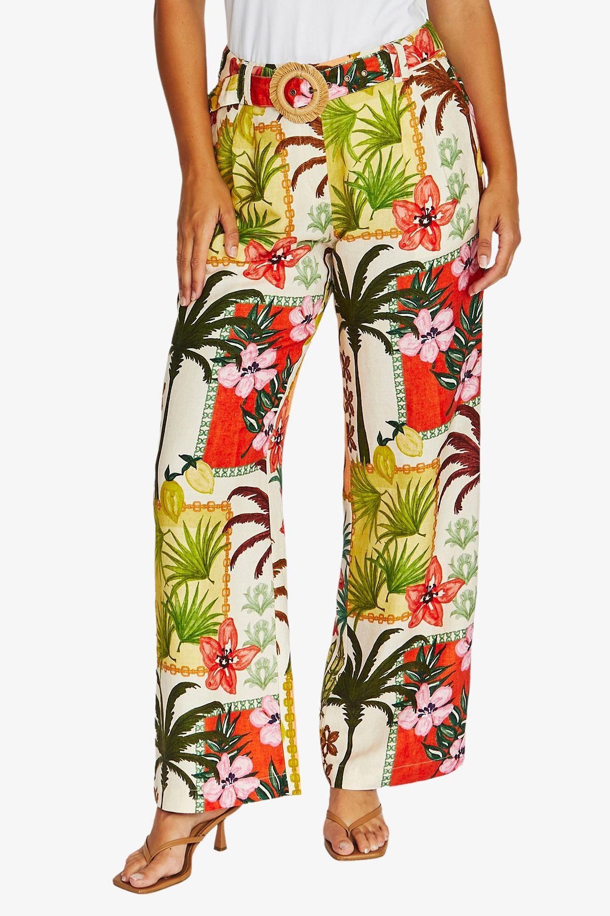 Front View high waisted, wide leg trousers with a vibrant tropical patchwork print featuring palm trees, hibiscus flowers, and red orange panels on a cream background. The pants are styled with a matching belt and brown heeled sandals.
