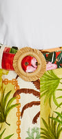 A close up of a tropical print belt with a large round woven straw buckle featuring a pink flamingo face, worn over a white top and paired with matching tropical print bottoms.