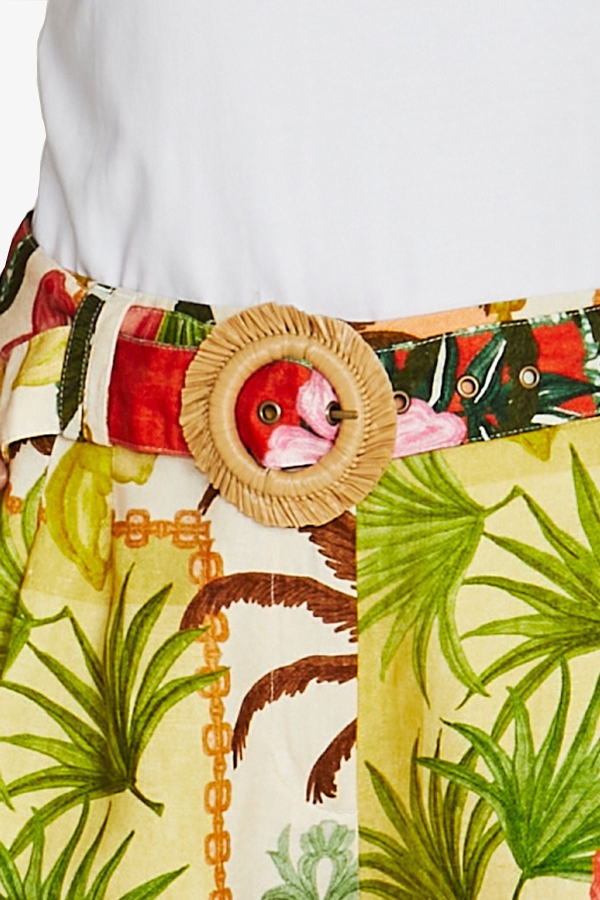 A close up of a tropical print belt with a large round woven straw buckle featuring a pink flamingo face, worn over a white top and paired with matching tropical print bottoms.
