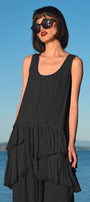 A model wearing a black longline silhouette top with a scoop neckline and a gathered, layered peplum hem, standing by a body of water with a blue sky in the background.