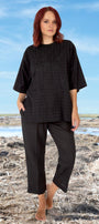A model wearing a black top and pants standing on a beach-like setting.