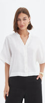 A woman modeling a white short-sleeve linen blouse with a split V-neck and black trousers, photographed against a clean white studio background.